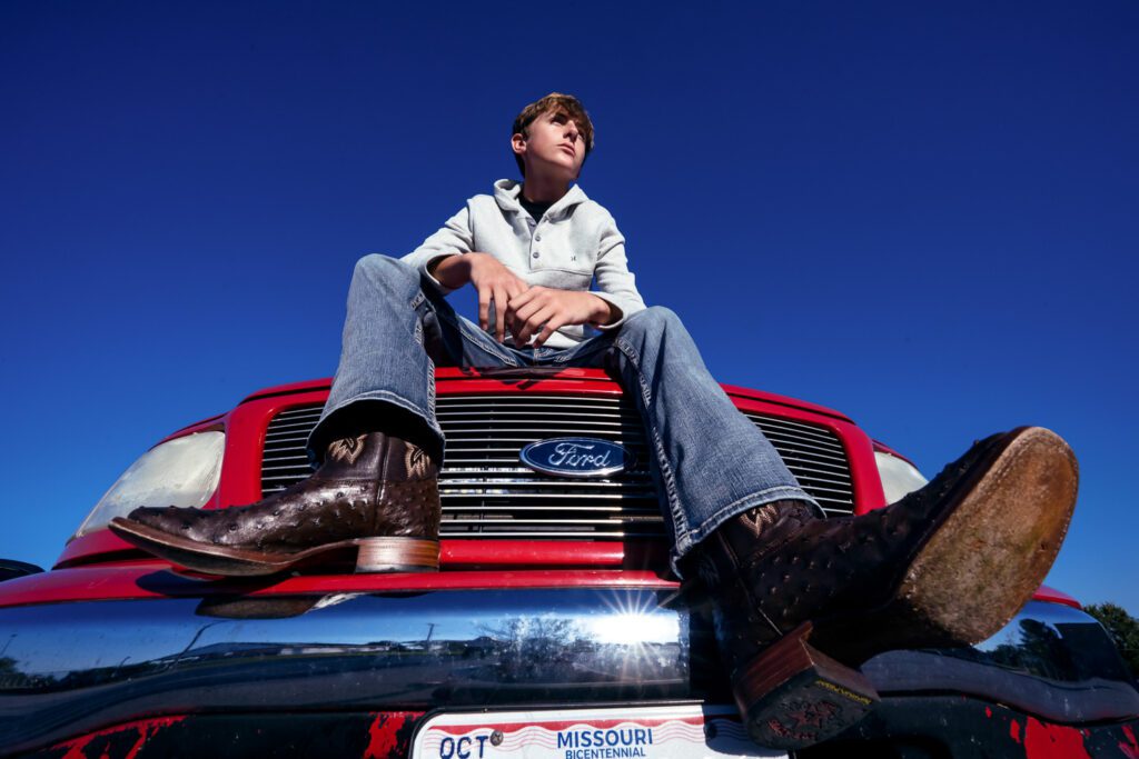 Senior guy sits on top of red truck with cowboy boots in foreground.