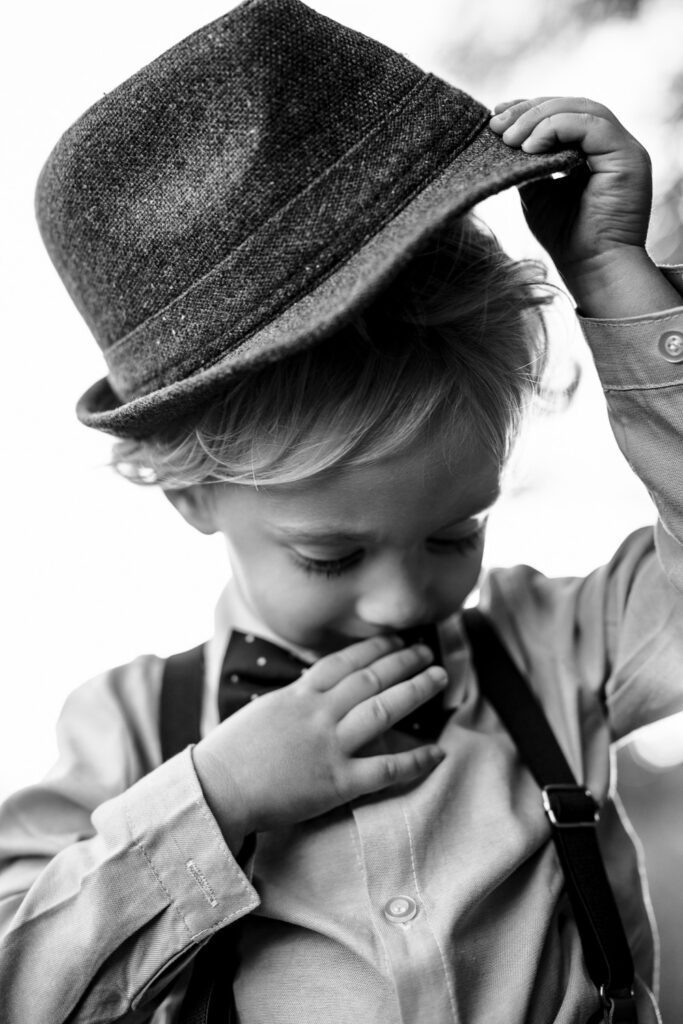 Young boy lifts his fedora hat while wearing suspenders.