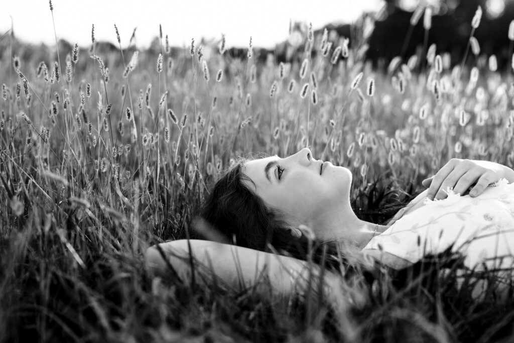 Senior girl with dark hair wearing white dress looks up at sky while laying in tall grass.