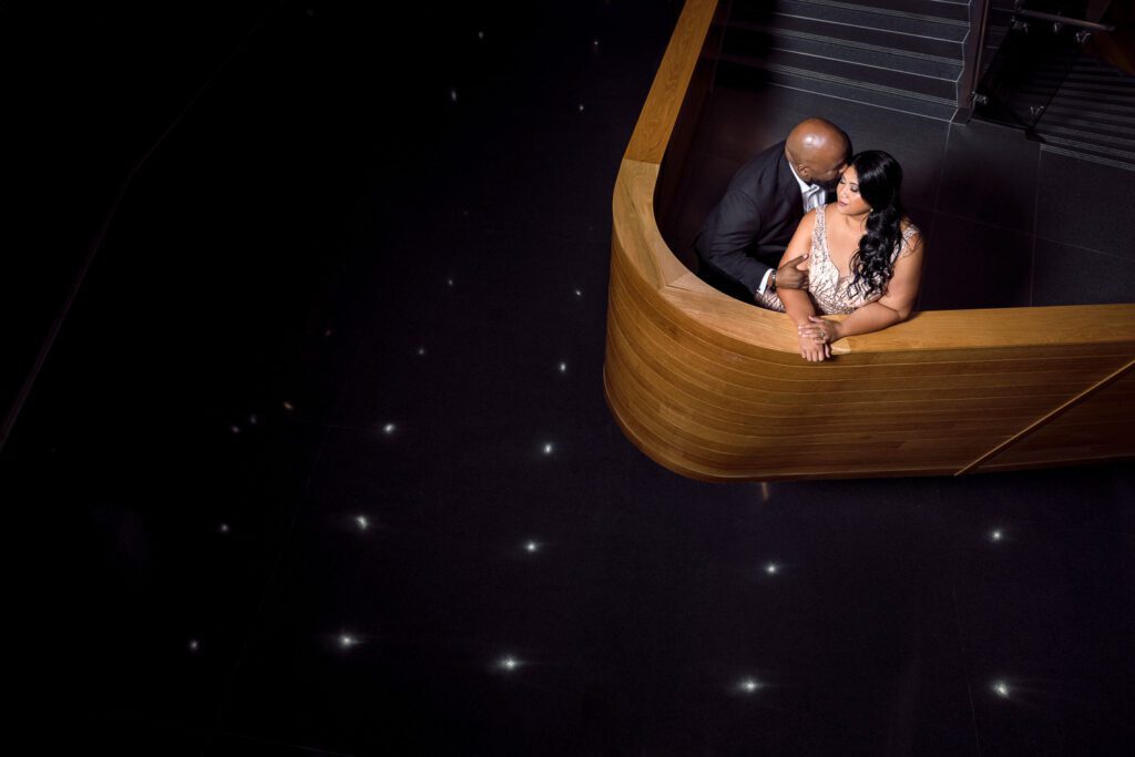 Couple pose on wooden stair case wearing formal attire while lights reflect on floor