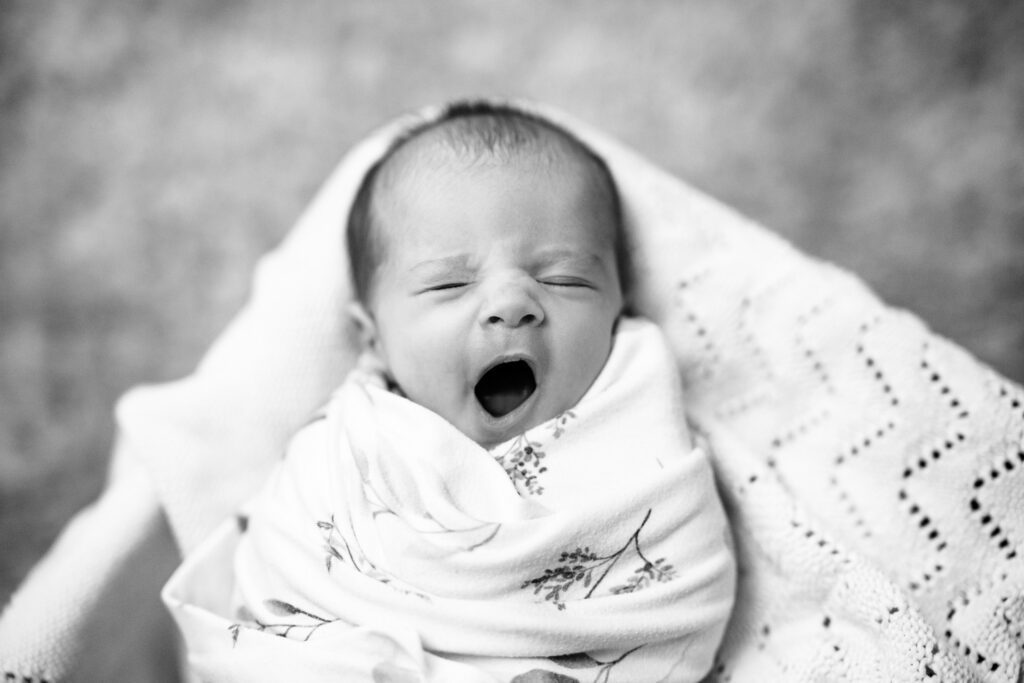 A black and white image of a newborn yawing while swaddleed in small box.