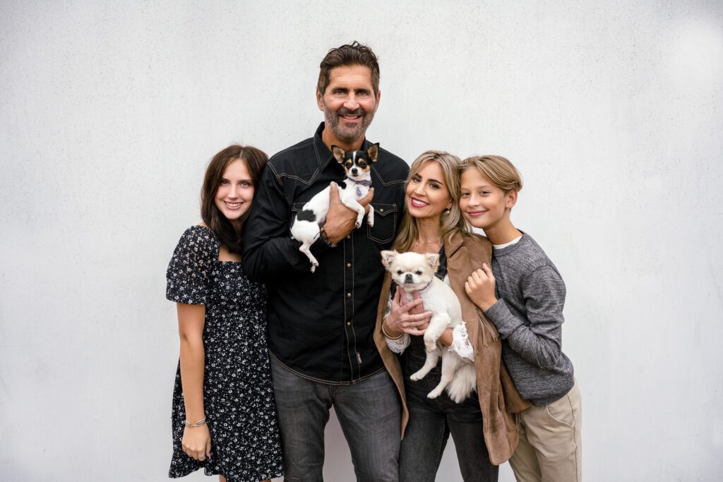 Family poses in front of white wall while holding two small dogs.