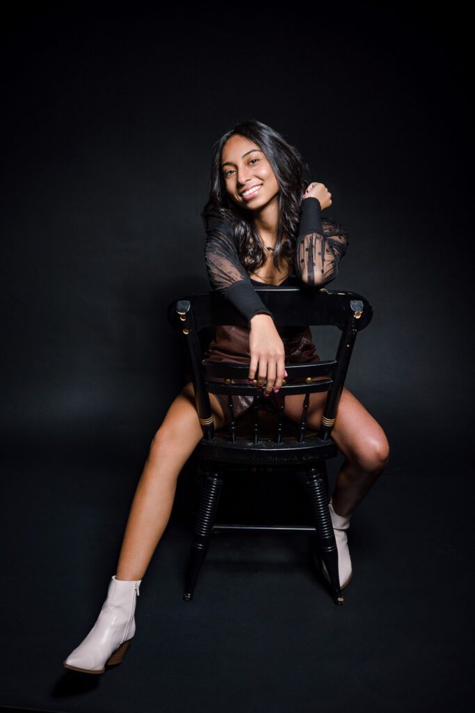 Senior girl poses on black backdrop while sitting in black chair wearing black lace top.