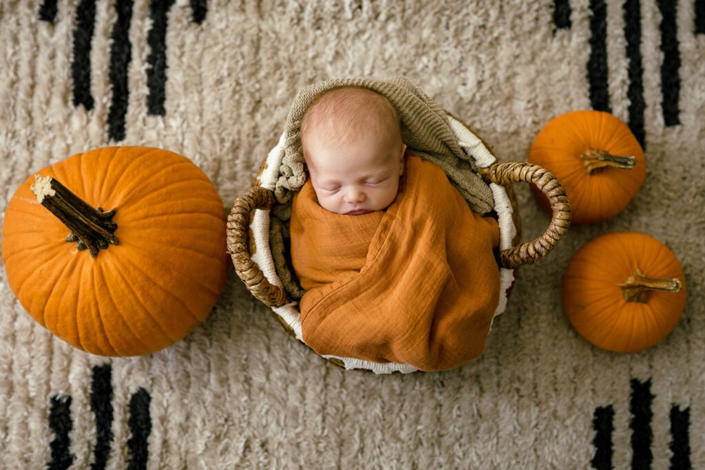 Newborn wrapped in orange swaddle surrounded by three orange pumpkins on beige rug.