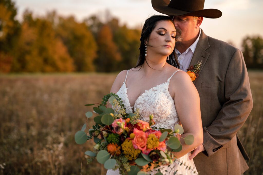 Couple kiss in fall field as wind blows on their wedding day. Groom is wearing a cowboy hat.