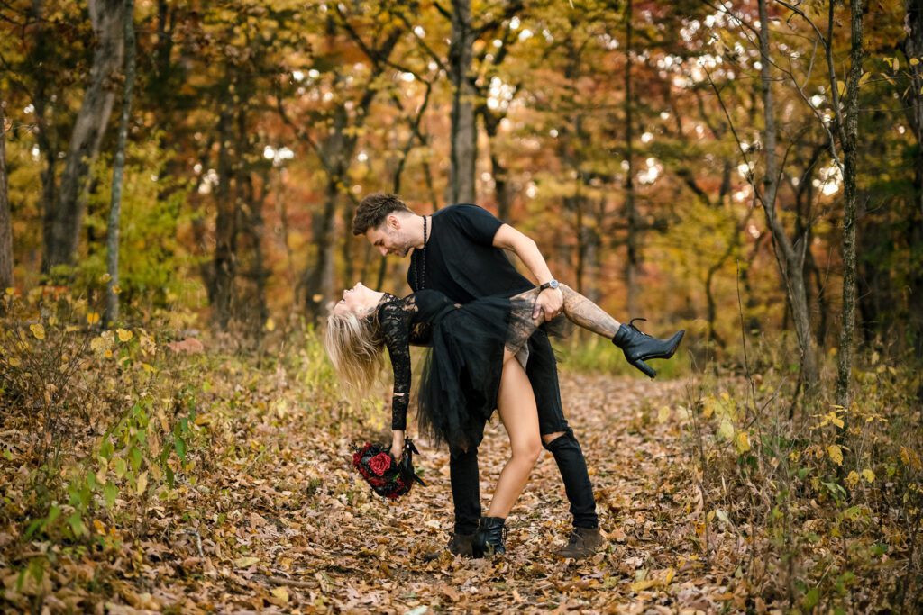 Hot couple wearing all black for Halloween in woods while husband dips wife while holding onto her.