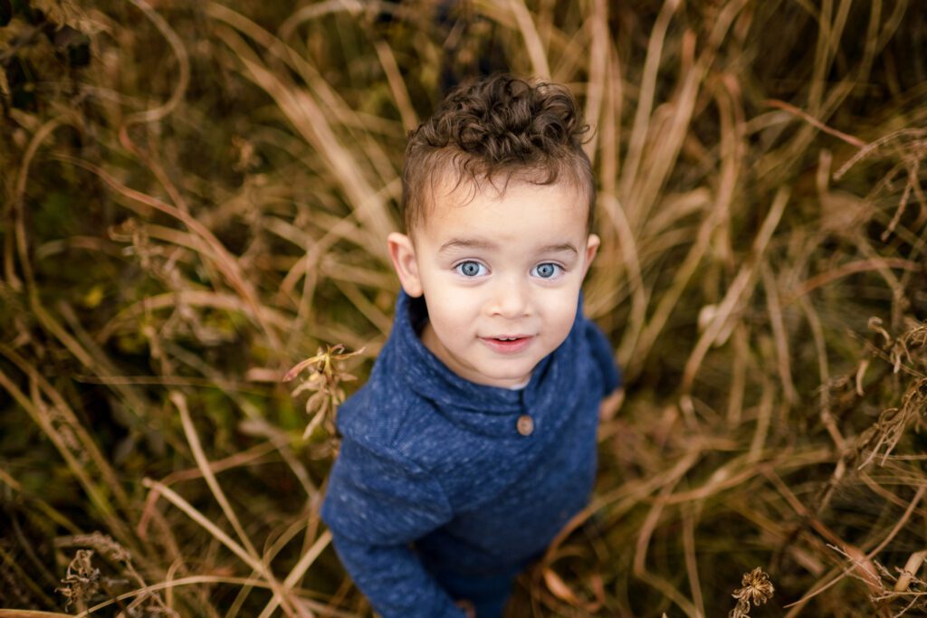 Young boy with blue eyes and blue sweater looks up at camera while standing in tall grass.