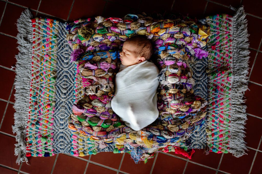 Newborn baby swaddled on colorful rug.