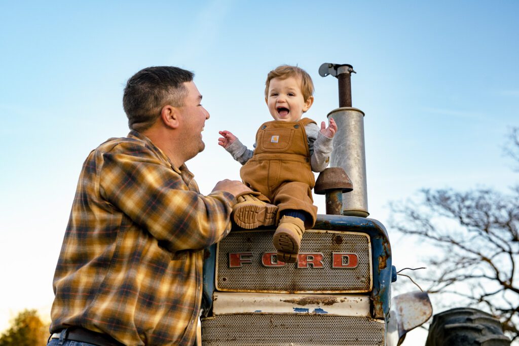 Young boy laughs at dad while sitting on top of old blue Ford Tractor.