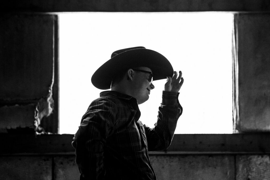 Black and White Image of senior guy tipping his cowboy hat in front of window.