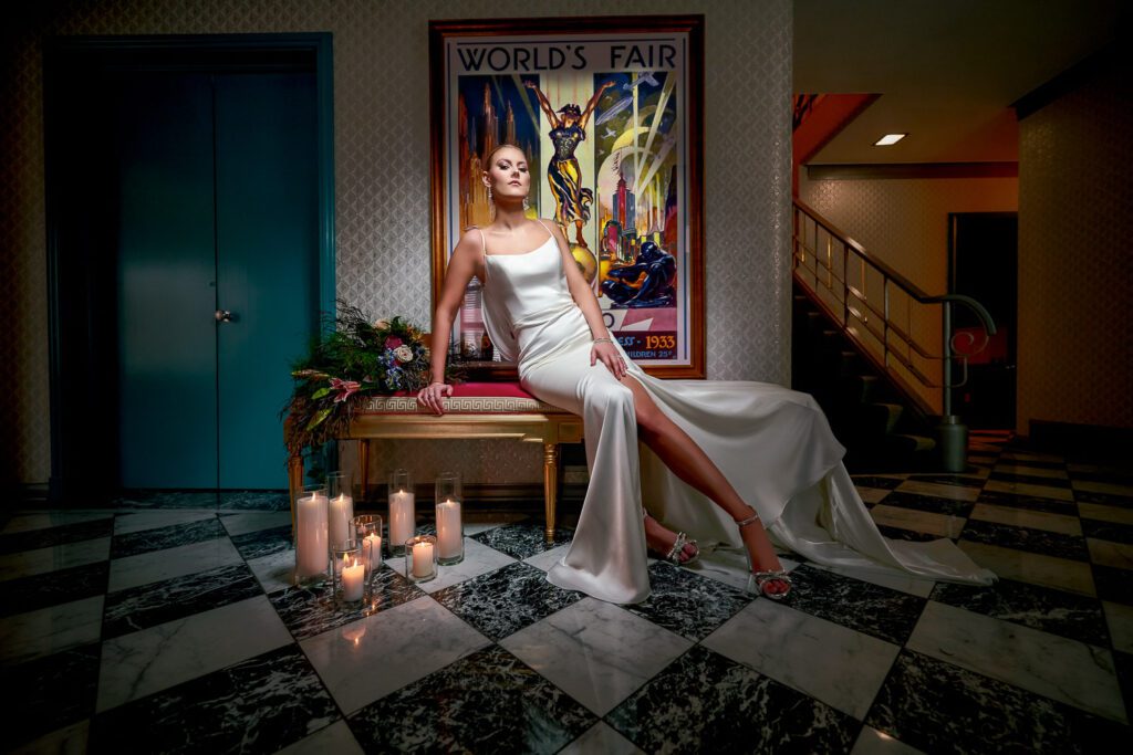 Bride in silk gown sitting on pink velvet bench with vintage worlds fair poster.