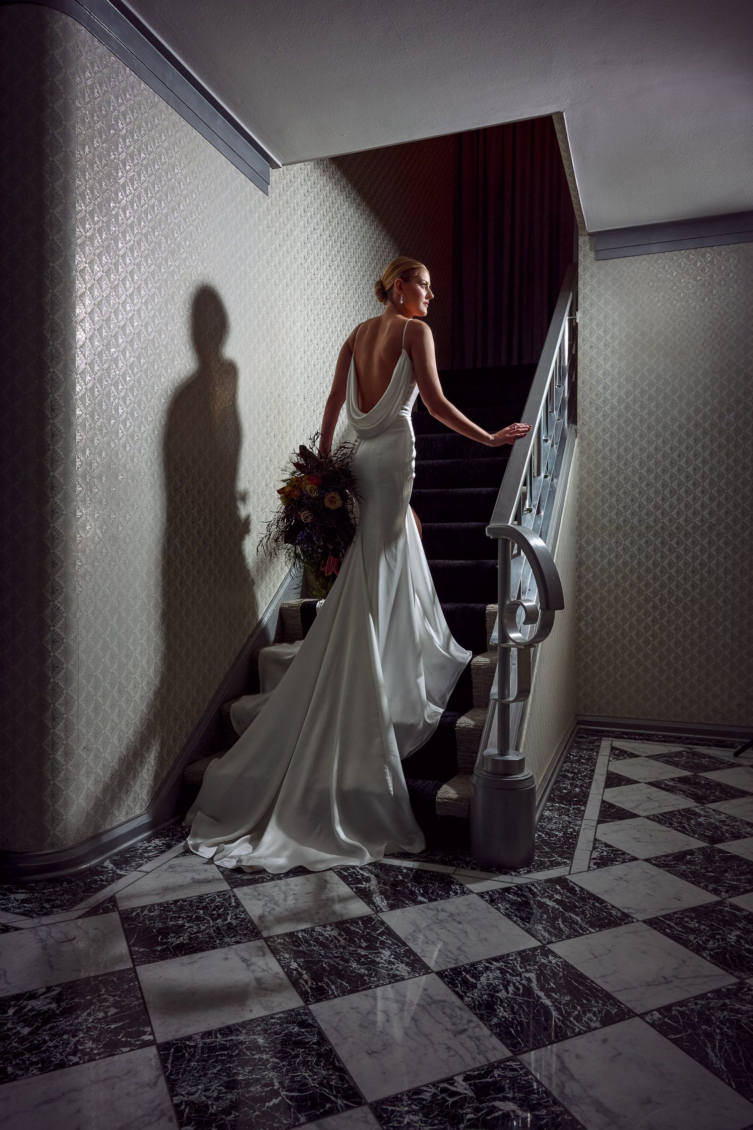 bride going up stairs in long silk cowl back gown with slicked back bun on marble checkerboard floors
