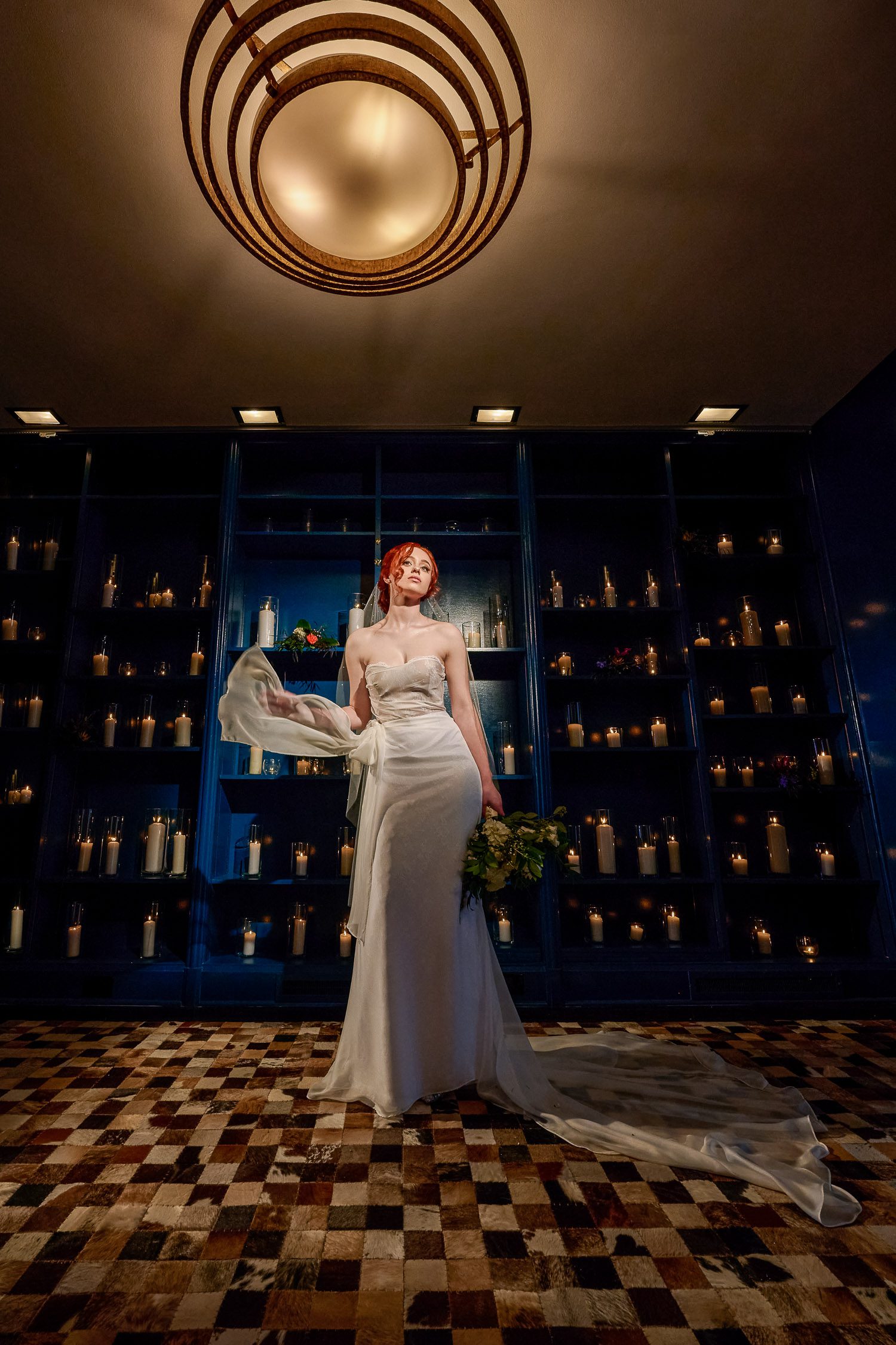 Bride wearing custom wedding gown stands in front of blue bookcase wall filled with candles at Chatol in Centralia, MO.