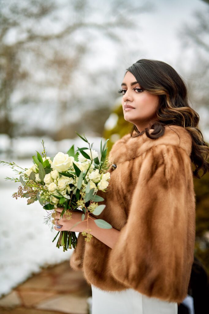 Indian bride looking off with fur cape and rose bouquet in snowy backdrop