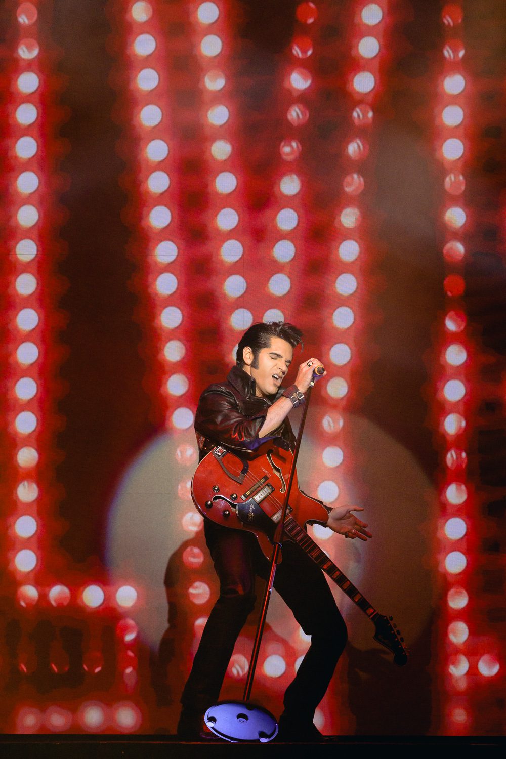 Dean Z singing into microphone wearing black leather with red guitar and red lights in background.