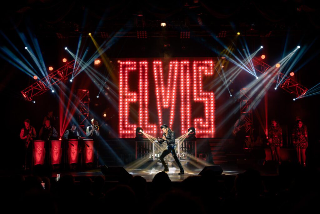 Dean Z under spot lights with Elvis in red letters wearing black leather suit singing by Schaefer Photography.