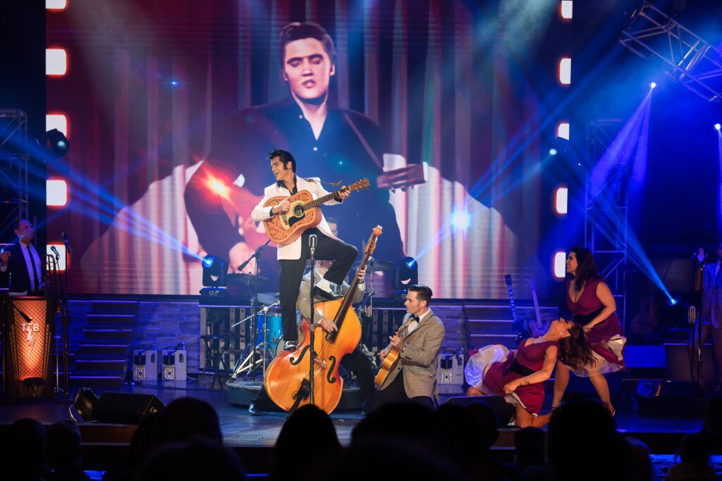 Elvis Presley Tribute Artist Dean Z dressed in 1950s suit standing on bass guitar as he plays an acoustic guitar.