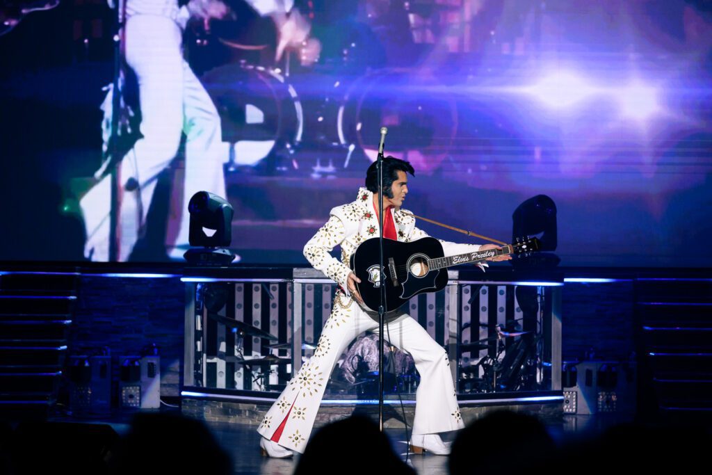 Elvis impersonator wearing white jump suit with red silk scarf pushes black guitar forward in a classic Elvis Presley Move.  Dean Z Ultimate Elvis performance by Schaefer Photography.