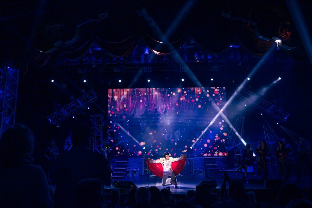 Dean Z takes a knee at the end of his performance as Elvis Presley while wearing a red cape that he is holding up in a traditional Elvis Presley manner.