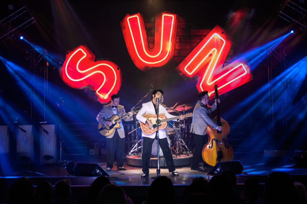 Dean Z with two musicians on stage un SUN records neon light projection.