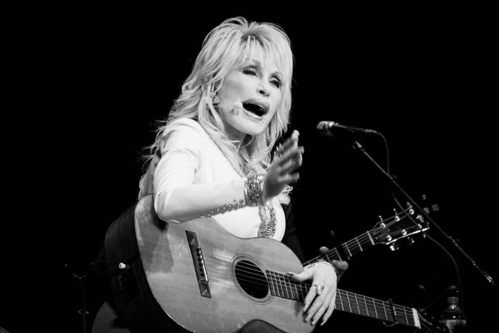 Dolly parton pionts with her hand while holding a guitar on her lap.