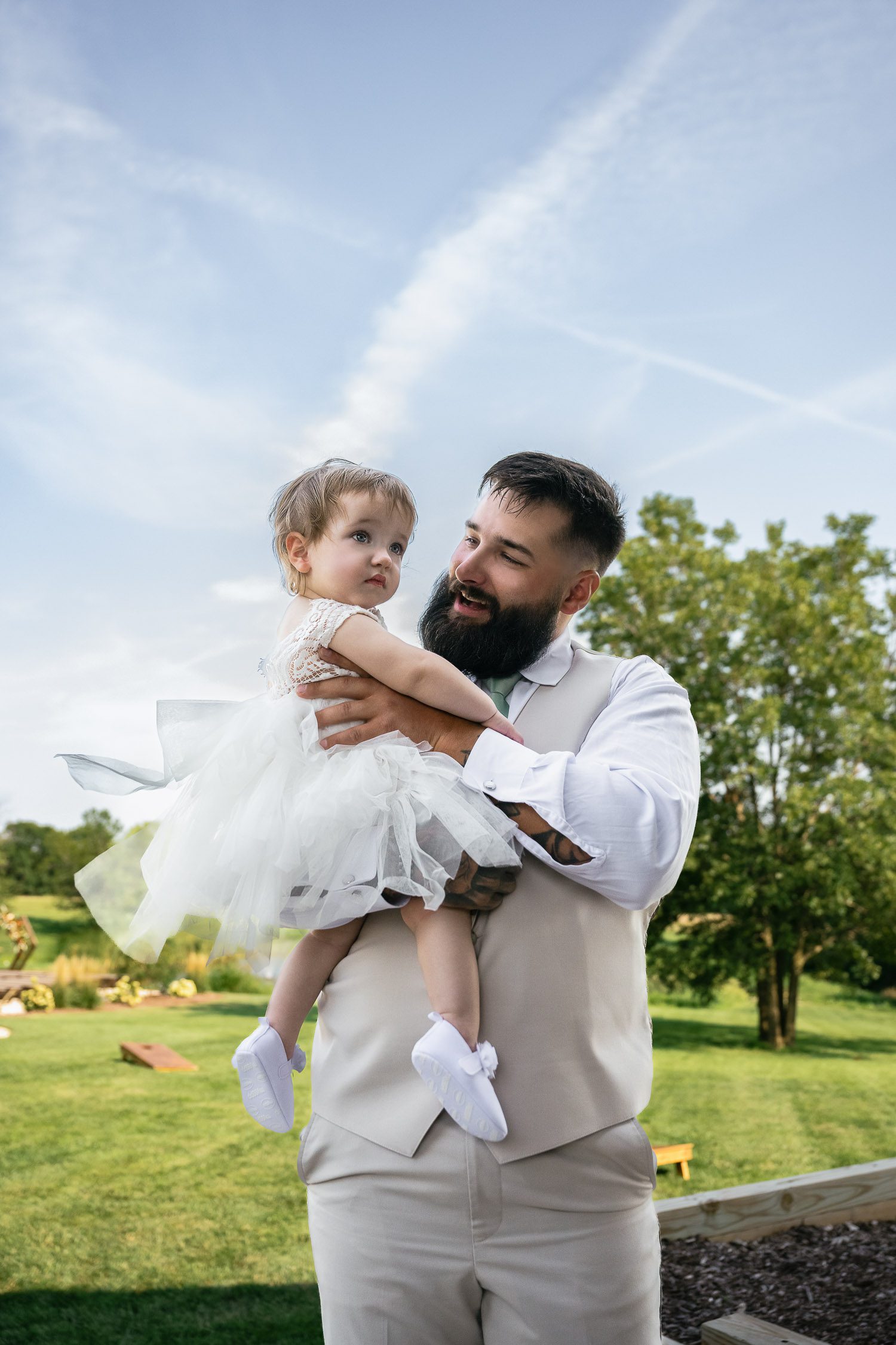 Groom holds daughter at wedding with blue sky and green tree