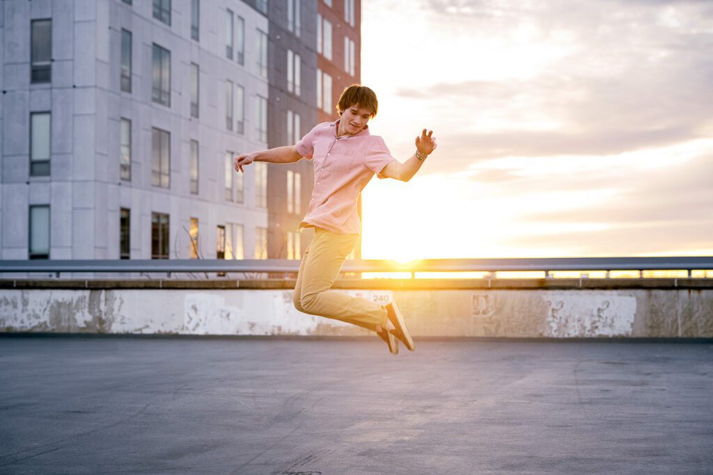 Senior guy jumps while kicking his heels in front of a setting sun and high rise buildings
