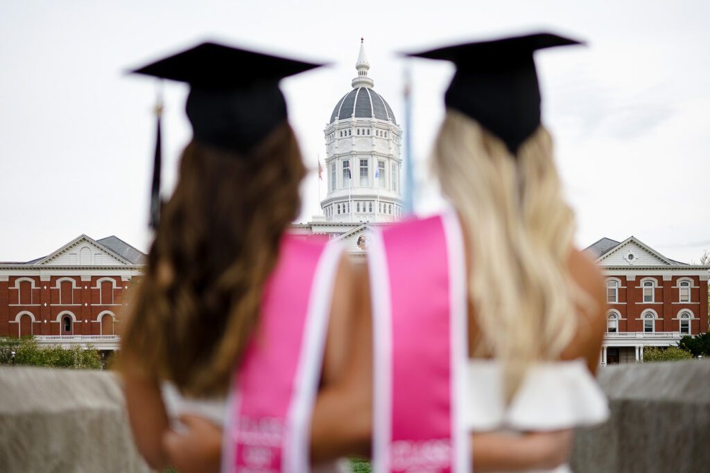 two women in graduation caps and pink sashes look at jesse hall mizzou