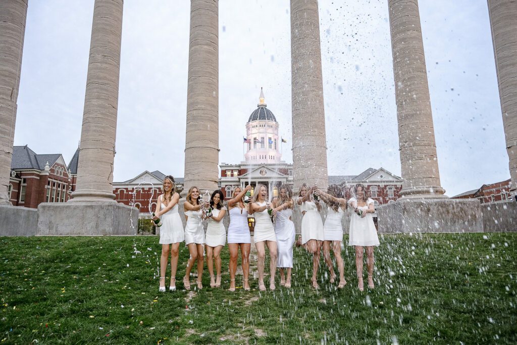 Sorority sisters wearing white dresses spray champagne in front of mizzou columns
