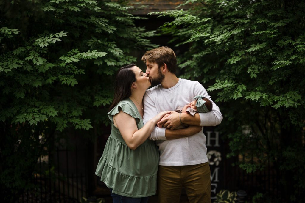 Couple kiss while holding newborn in front of green leaves