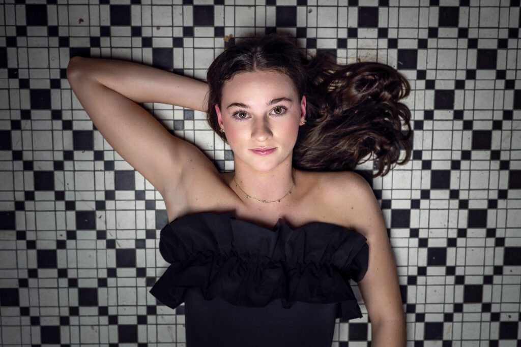 Senior girl with black dress lays on black and white tile flooring