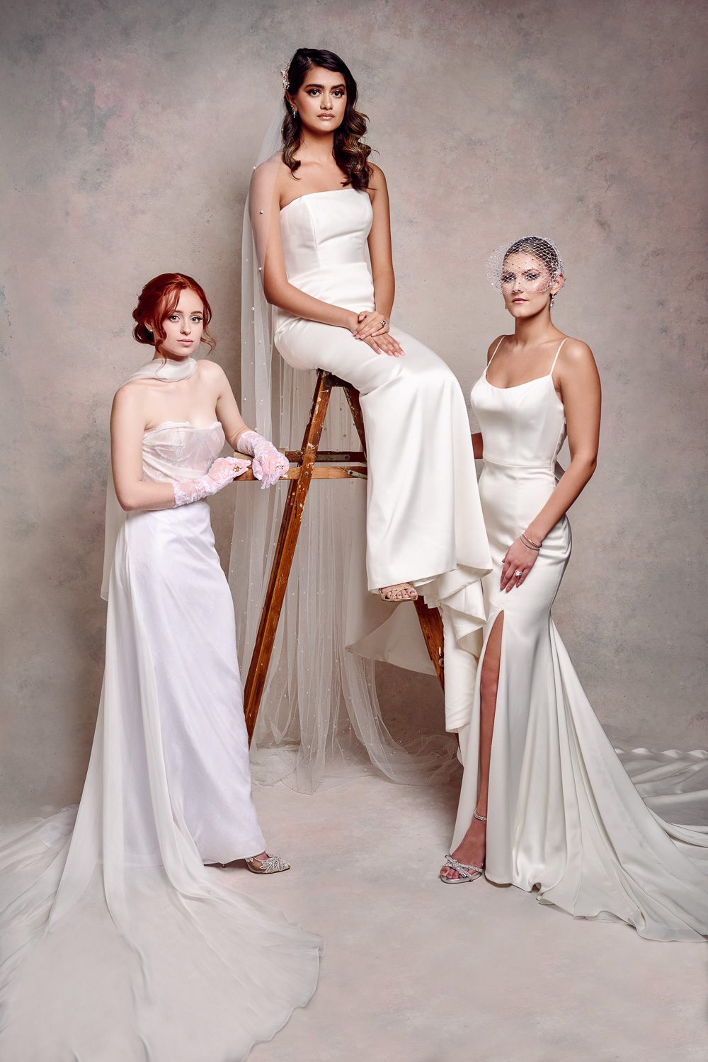 Three models wearing wedding dresses pose on ladder with painted canvas backdrop