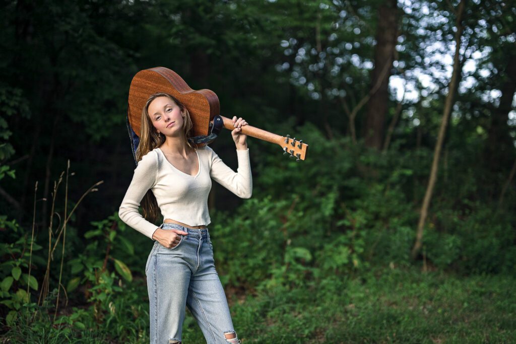 senior girl with white top and blue jeans holds acoustic guitar on shoulder