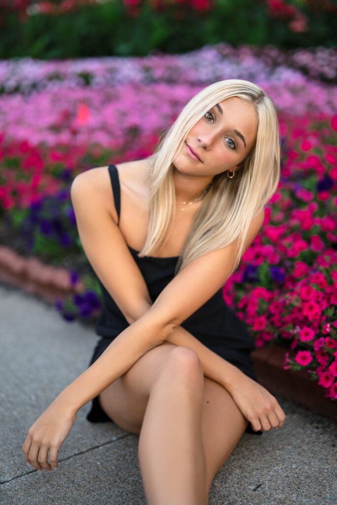 Beautiful blonde senior girl sitting in front of pink flowers wearing black dress