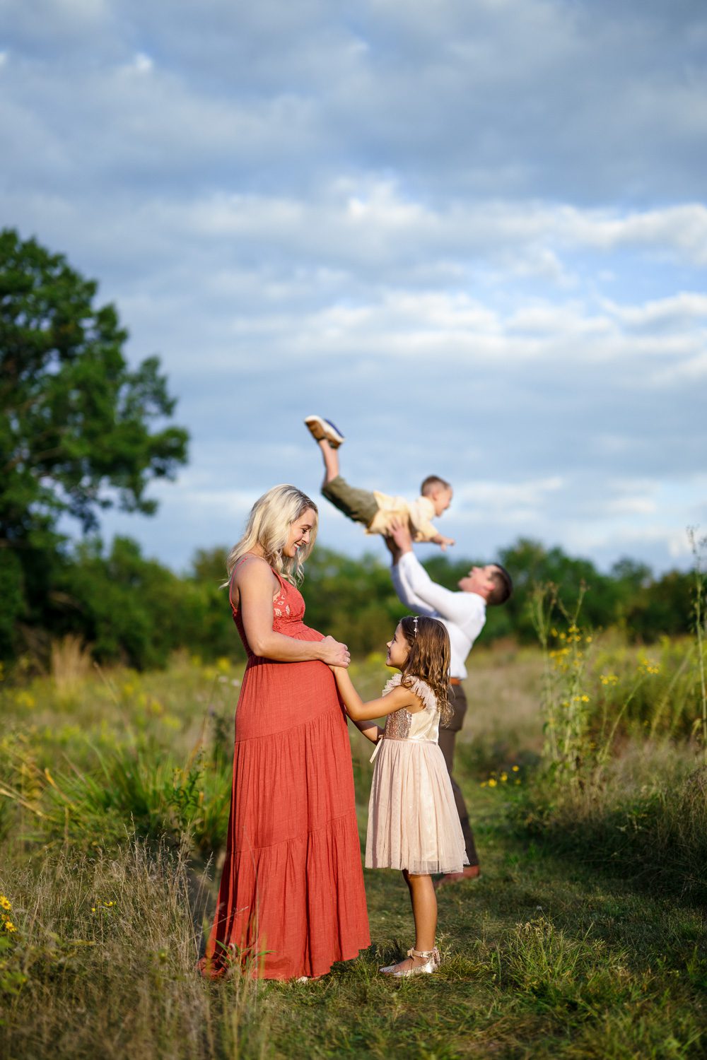 Expecting mother holds daughters hands while father and son play in back 