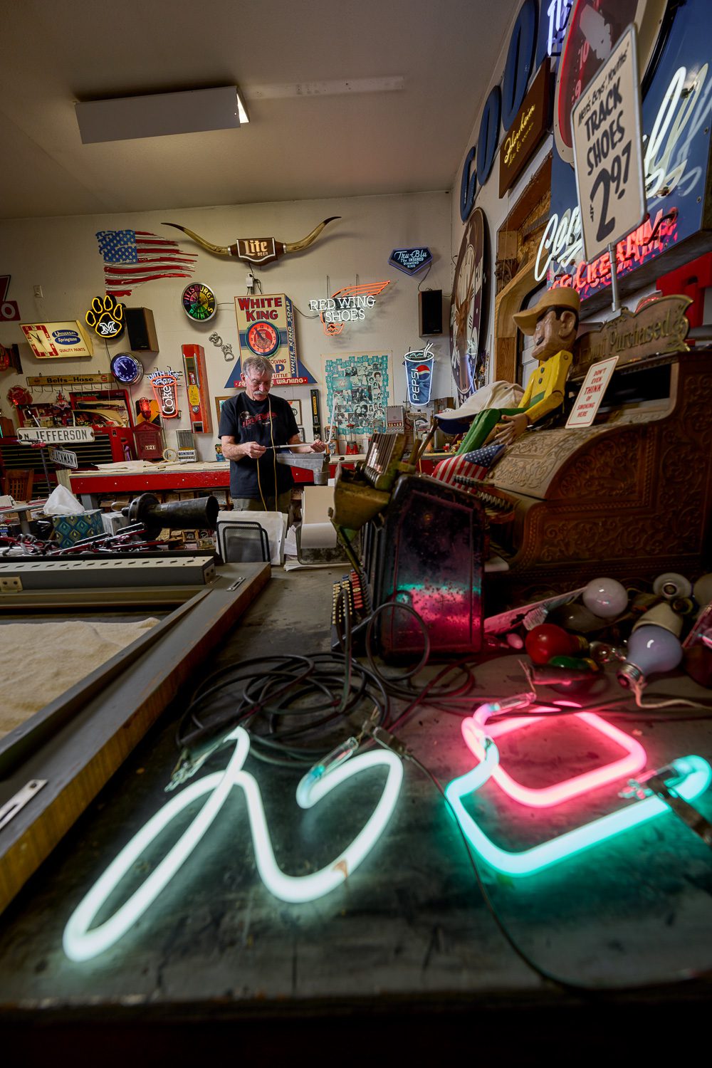 Jim McCarter working in his work shop with neon signs part burning in foreground.