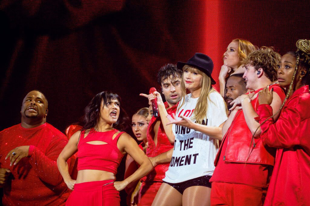 Taylor swift wearing black hat and white tshirt with writing with dancers wearing red