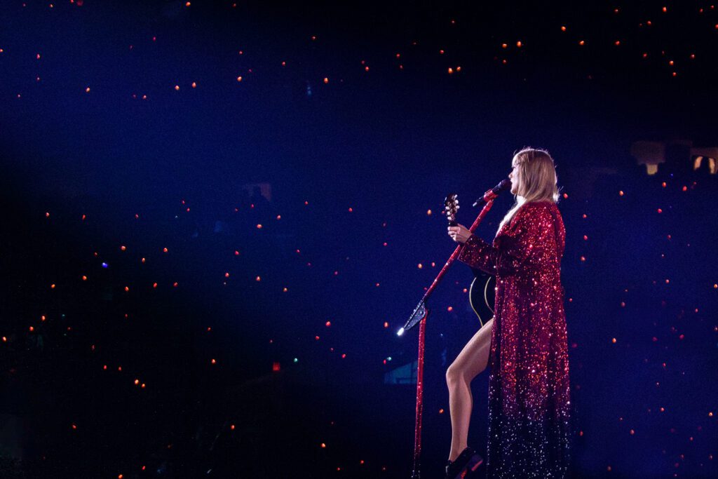 a spotlight shines on taylor swift wearing red sequin floor length jacket with guitar