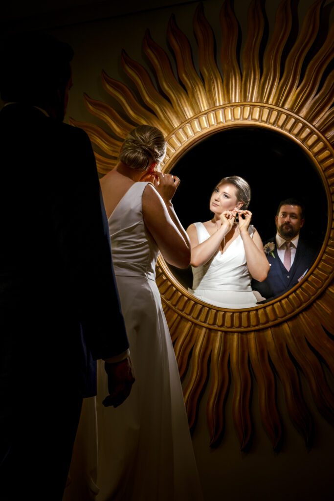 Bride Adjusts earring while looking into gold circle mirror with groom