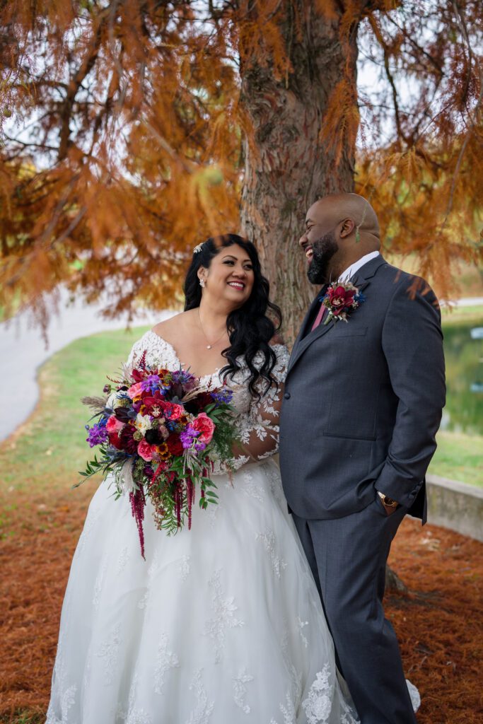 bride and groom stand under rust color tree in fall during wedding photos
